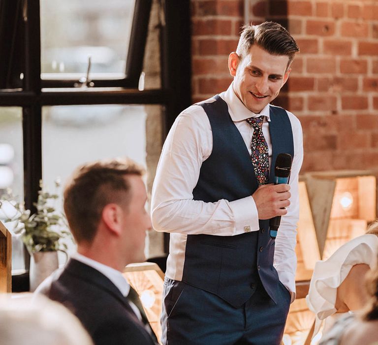 Groom in navy blue waistcoat stands as he reads out his wedding speech and looks at guest 