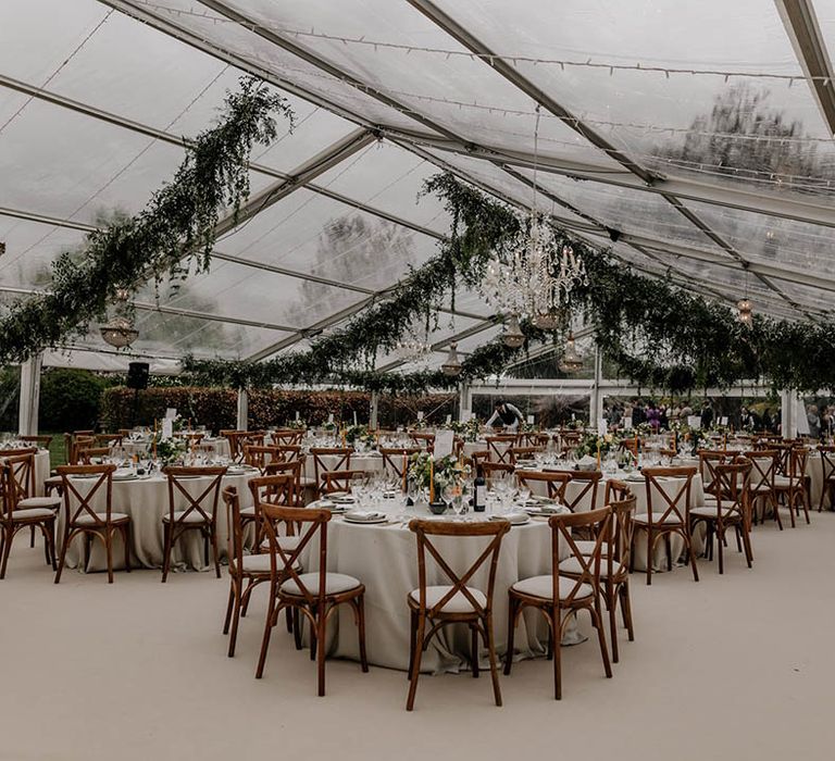 Clear glass marquee wedding reception with wooden furniture and hanging greenery garlands 