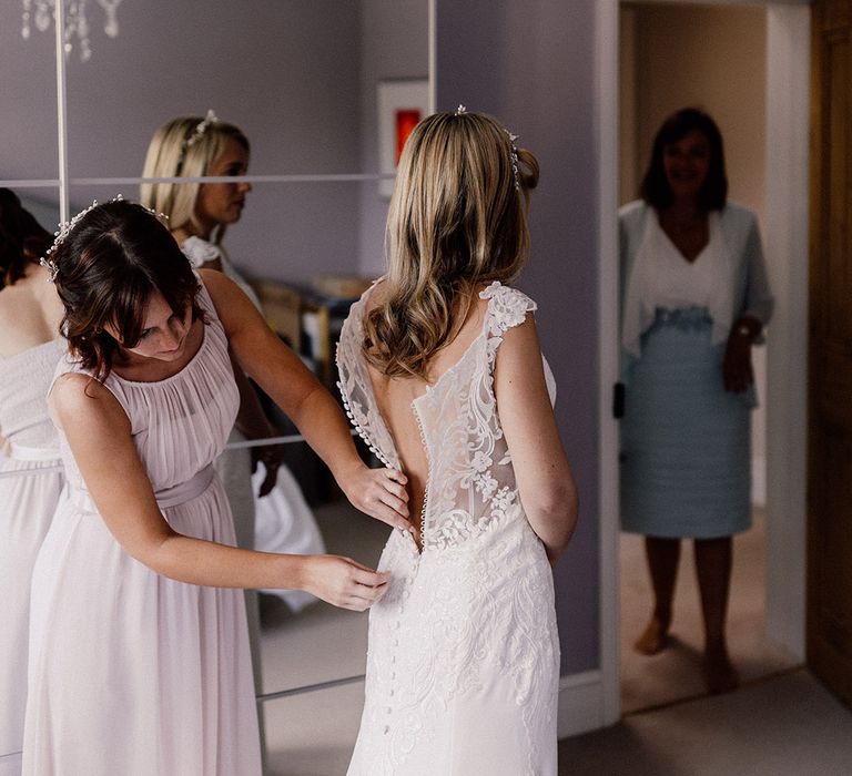 Bridesmaid in blush pink chiffon dress buttons up the bride into her lace wedding dress 