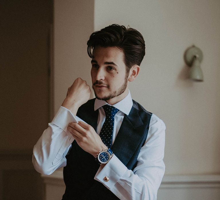 Groom fixes his cufflinks wearing a blue checkered waistcoat and trousers with a polka dot blue and white tie 