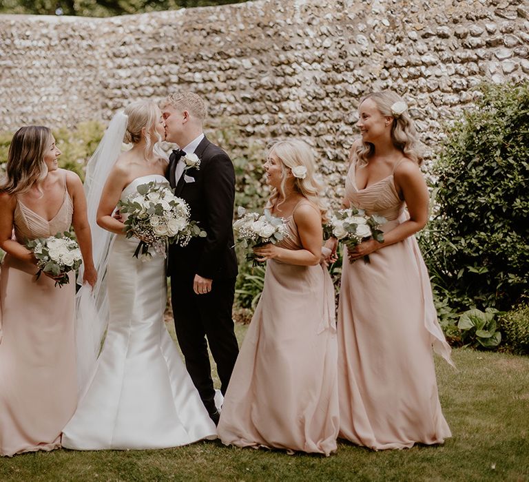 Bride and groom kiss as the bridesmaids turn to look in their champagne pink dresses 