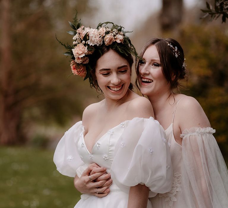 Bride in a floaty wedding dress with detachable sleeves embracing her partner in a strapless wedding dress with puff sleeves and flower crown 