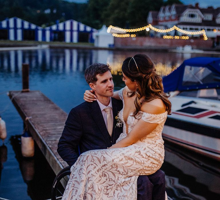 Groom in a blue suit with a gold tie with the bride sitting on his lap wearing an off the shoulder boho lace fitted wedding dress and gold head piece 