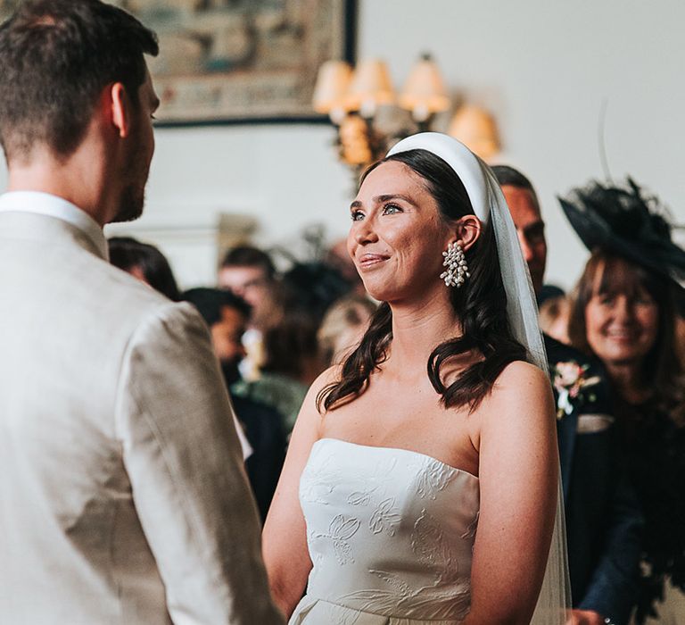 Bride in Jesus Peiro headband and pearl statement earrings smiles at the groom during their ceremony 