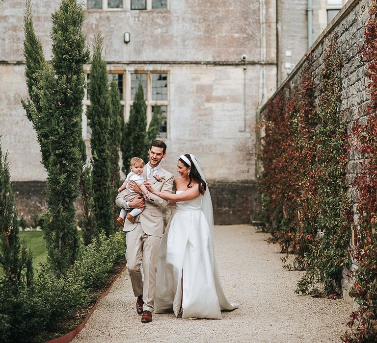 Bride and groom walk around the grounds of Elmore Court with their baby boy