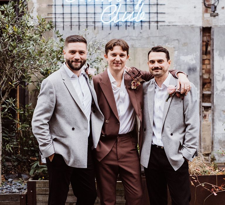 Groom in brown suit with unbuttoned shirt with the best men in grey suits and pink flower buttonholes