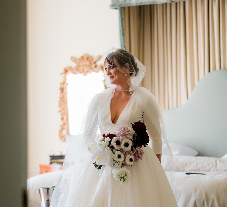 Bride wears hood styled veil with tulle skirt princess dress with V-shaped neckline whilst holding bright floral bouquet 