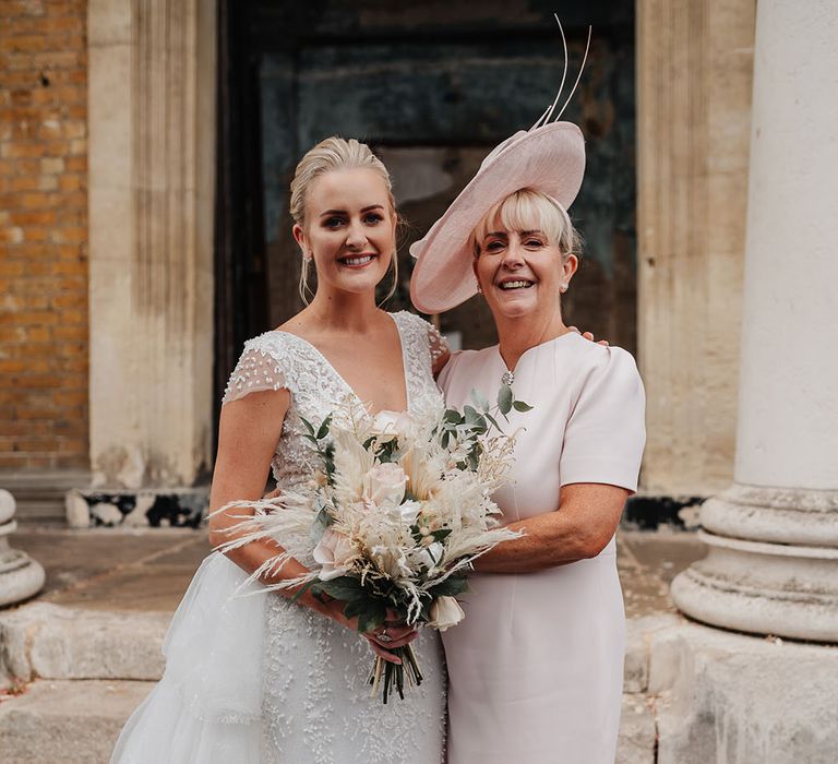 Bride holds pampas grass bouquet and stands with her mother who wears pale pink dress and fascinator 