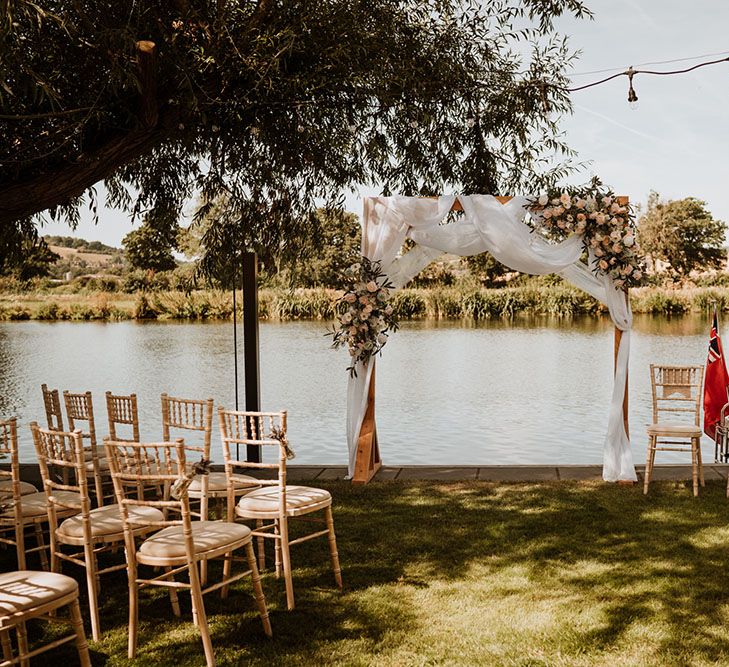 Wooden arch complete with white fabric draped across and floral decor in front of wooden seats for outdoor ceremony beside the Thames