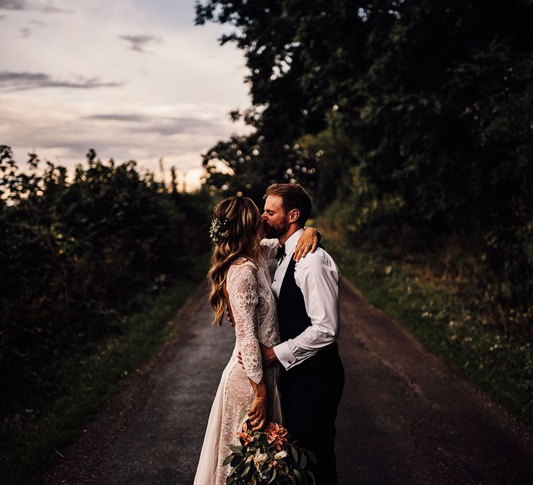 Groom kisses the bride in a lace wedding dress holding a yellow and white wedding bouquet 