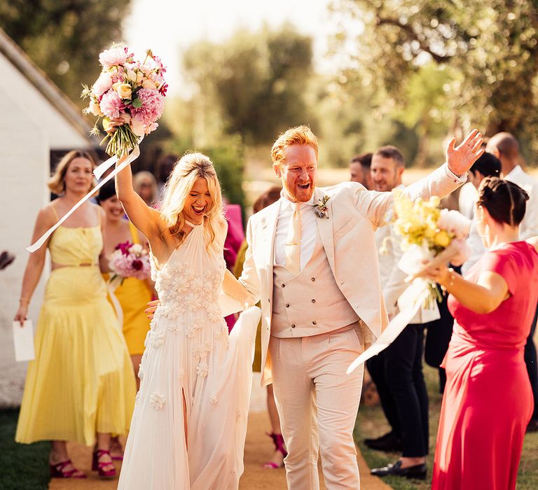 Groom in a cream suit walking with the bride in a halter flower Emma Beaumont wedding dress 