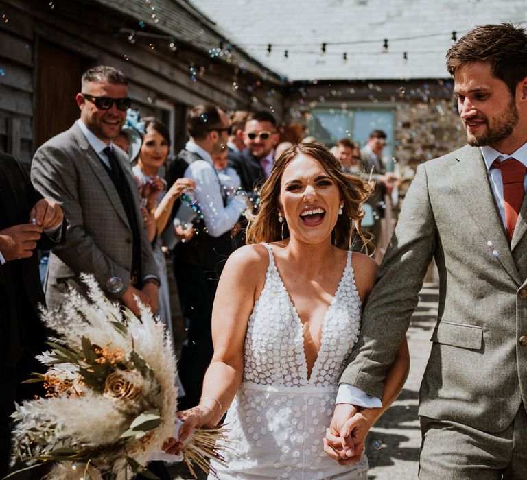 Bride in sleeveless Made With Love Bridal dress with white embellishments and deep plunge neckline and groom in grey suit with orange tie doing confetti walk at The Green Cornwall 