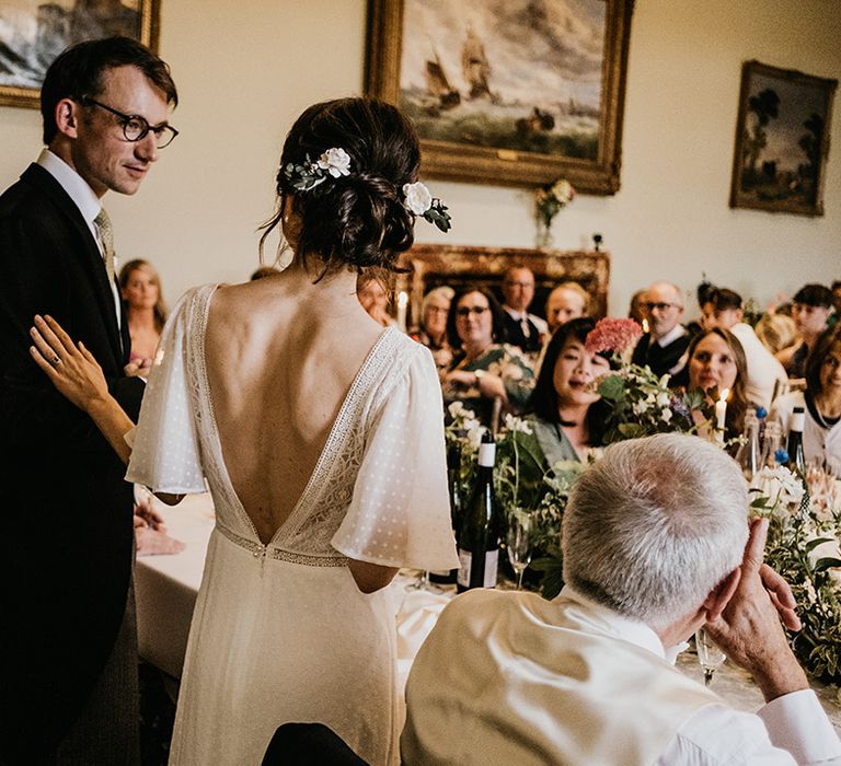 Bride in open back wedding dress with batwing sleeves and polka dot details with white rose hair clip and groom in dark three piece suit