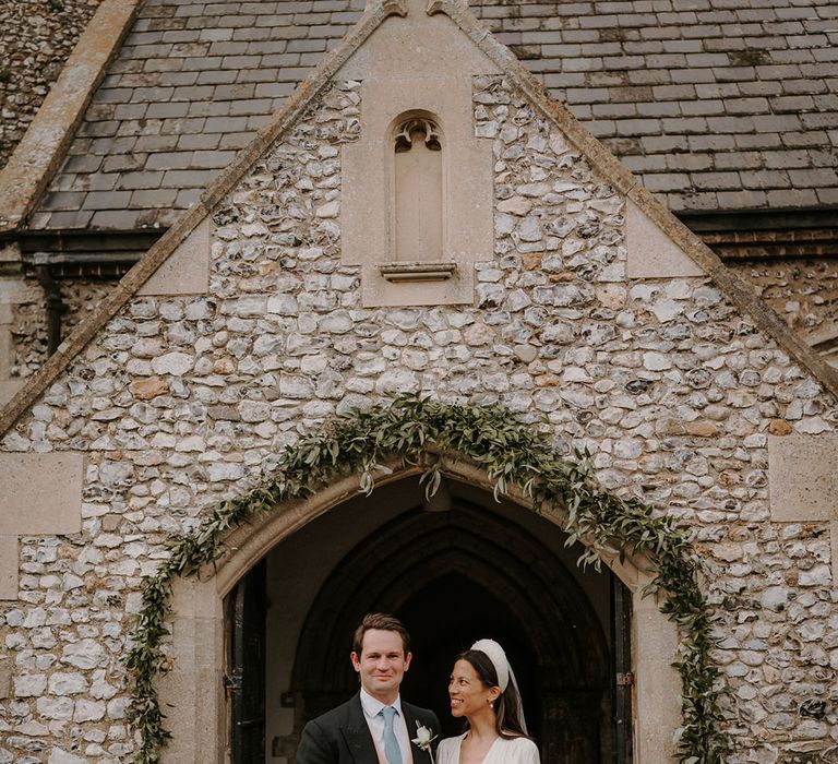 Groom in a morning suit with a pink waistcoat and blue tie with the bride in a satin wedding dress standing at the entrance to the church