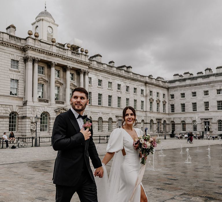 Bride in modern v neck puff sleeve wedding dress with front slit and groom in dark suit walking around Somerset House