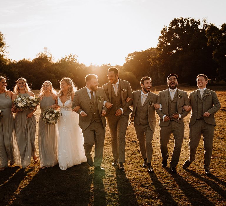 The bridal party and the groomsmen walk with the bride and groom during golden hour laughing together 