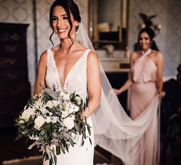 Bride wearing pearl drop earrings holding a white flower bouquet stands smiling with a bridesmaid in a pink dress holding her train 