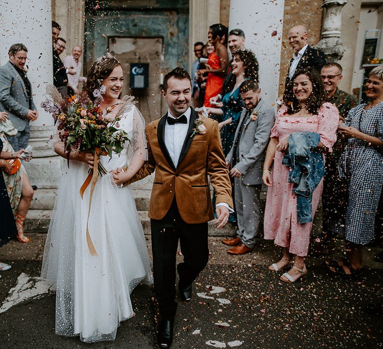 Bride and Groom walk through confetti throw wearing a coloured suit jacket and celestial wedding dress whilst carrying an autumnal bouquet full of Thistles, dried grass and Lila Delphiniums