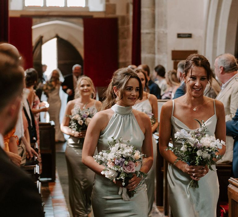 Bridesmaids in sage green satin dresses with different styles walking down the aisle holding small pastel bouquets 