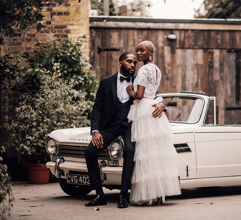 Black Bride and Groom pose in front of classic vintage wedding car whilst wearing a black suit and a bridal two piece set