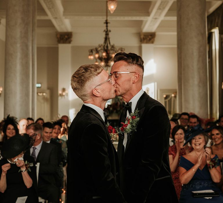 Grooms in matching velvet blazers with white shirts and black bowties with red rose and foliage boutonniere kissing at The Balmoral Edinburgh