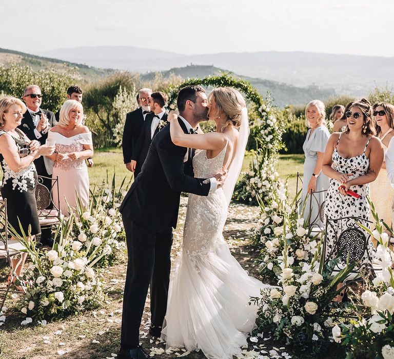 Bride in lace wedding dress kisses her groom in front of floral archway at the Castello di Petrata Umbria Villa  