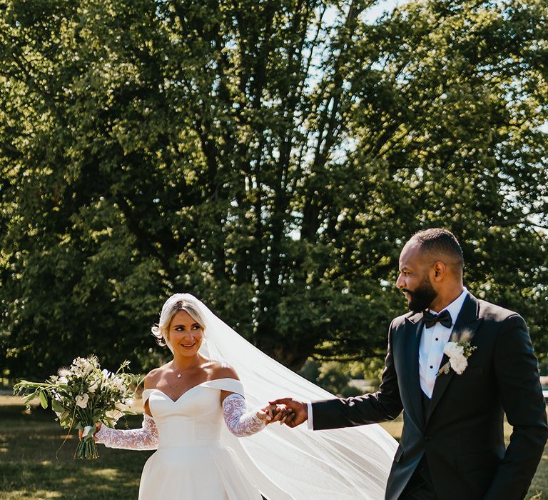 Bride in off-the-shoulder princess wedding dress with detachable lace sleeves walks alongside her groom in black-tie