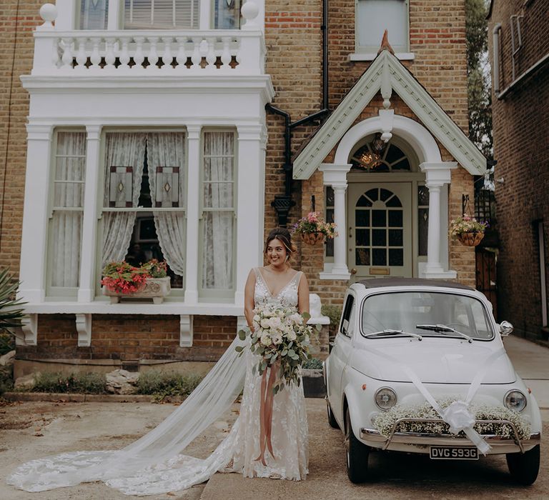 Bride in sleeveless plunge lace wedding dress  and cathedral length embroidered veil standing beside a classic fiat 500 wedding car 