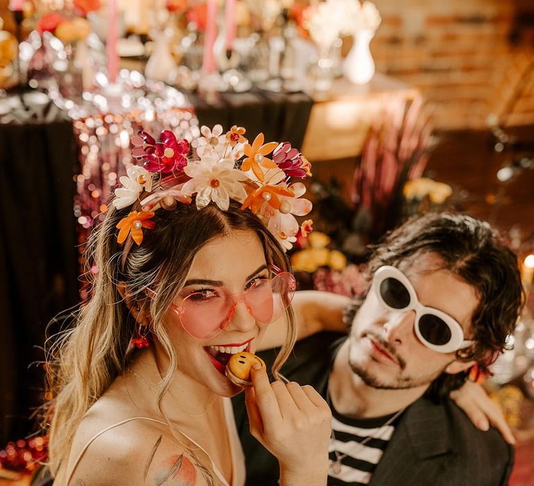 Bride in v-neck satin wedding dress and artistic flower crown eating a cookie with groom in dark grey pinstripe blazer and black and white long sleeve t shirt and large white sunglasses 