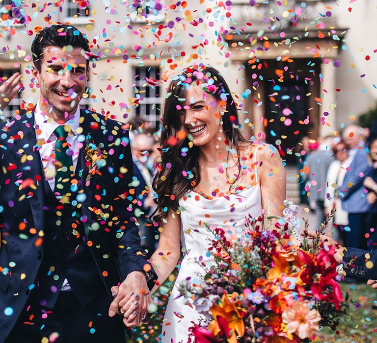 Groom in three piece navy suit with green tie standing with bride in satin wedding gown walking out to colourful confetti 