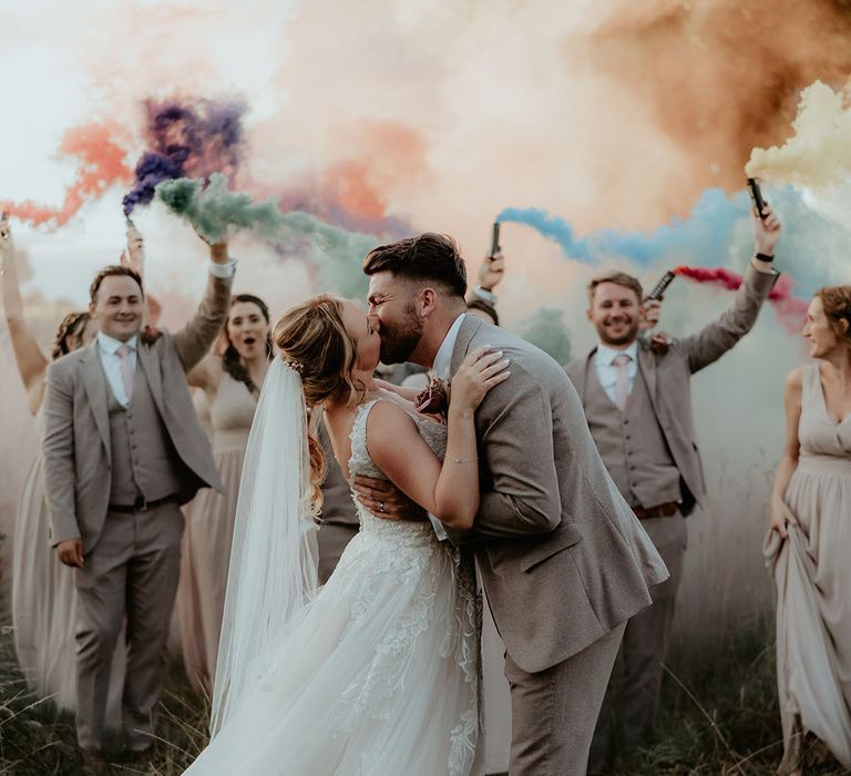 Bride & groom kiss surrounded by their wedding party holding colourful smoke-bombs 