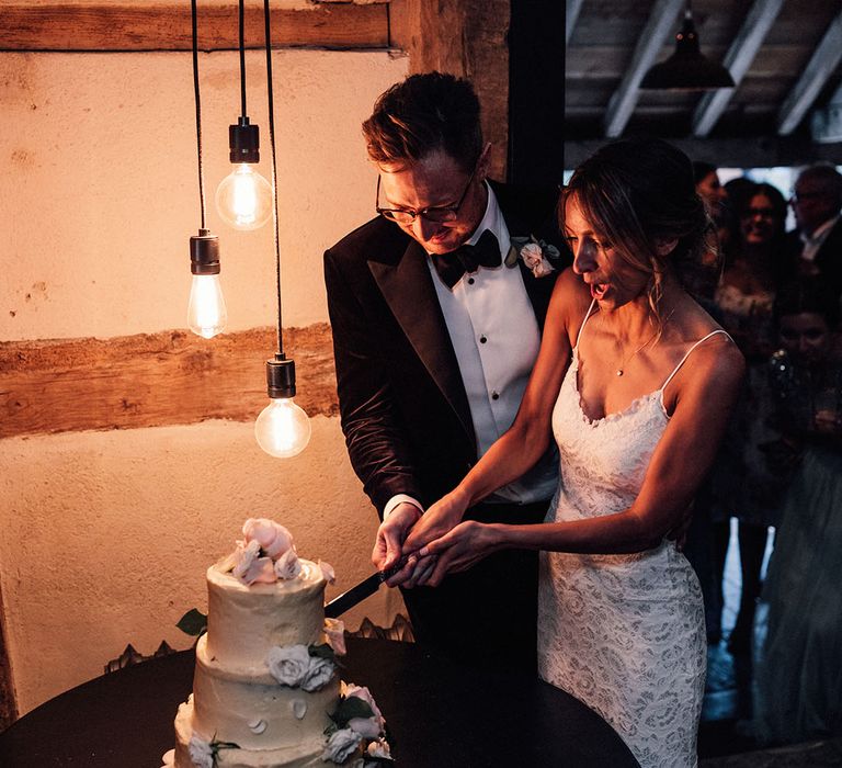 Three tier buttercream frosted wedding cake with pink rose petal decor being cut by the bride and groom with cosy festoon lighting 