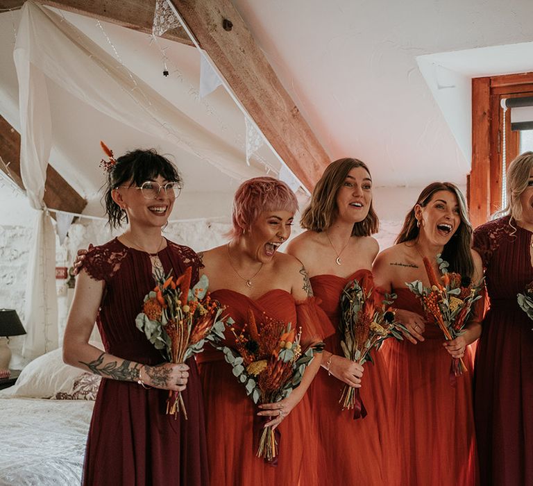 Mismatched burgundy and burnt orange bridesmaid dresses holding dried flower and eucalyptus bouquets doing first look with bride 