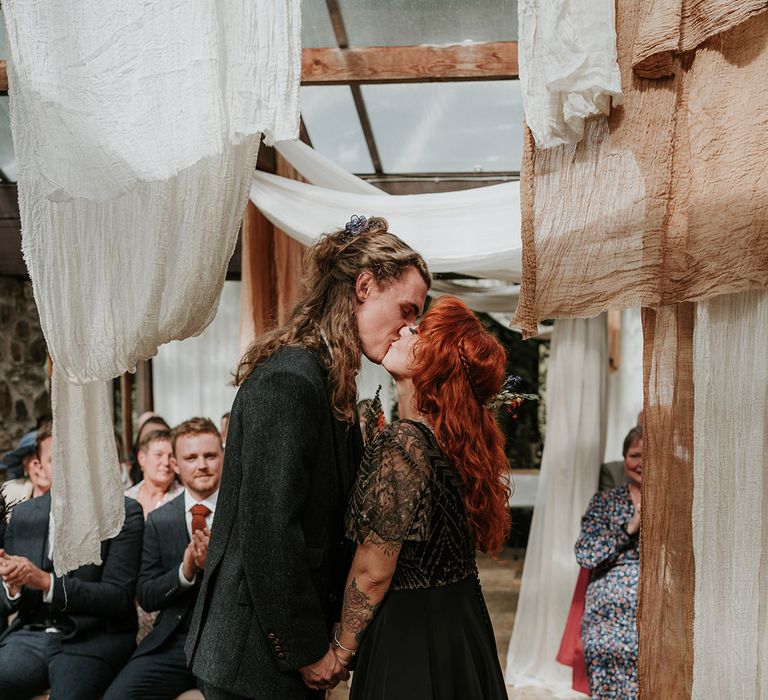 Groom in dark grey three piece suit and burgundy flower patterned tie kissing bride in embellished black wedding dress with sheer sleeves standing in Westcott Barton wedding venue with off-white and orange ceiling drapes 