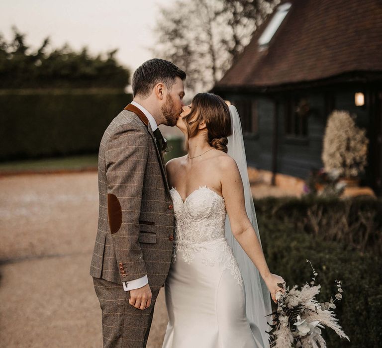 The groom in a grey checkered suit kisses the bride in a strapless satin finish wedding dress 