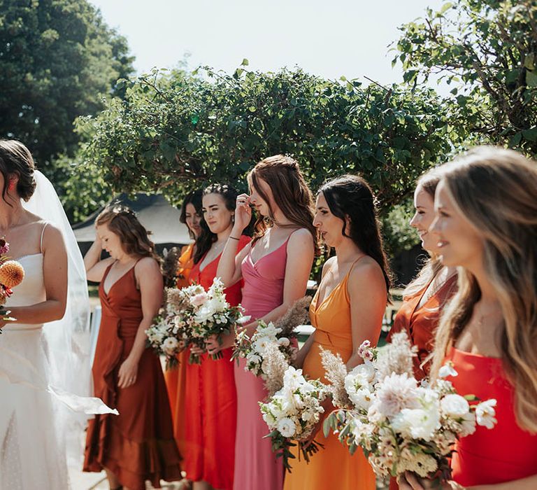 Bride in a strappy Stephanie Allin bridal gown holding a pompom dahlia bouquet and bridesmaids in mismatched bridesmaid dresses