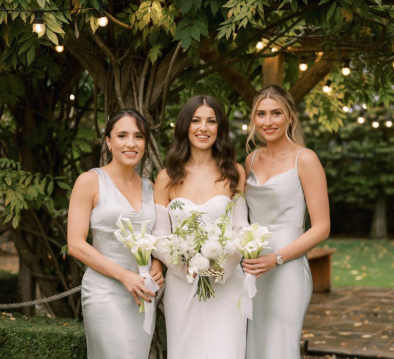 Bridesmaids in mismatched sage green dresses with the bride in a strapless corset wedding dress alll holding with flower bouquets with lilies, dahlias and more