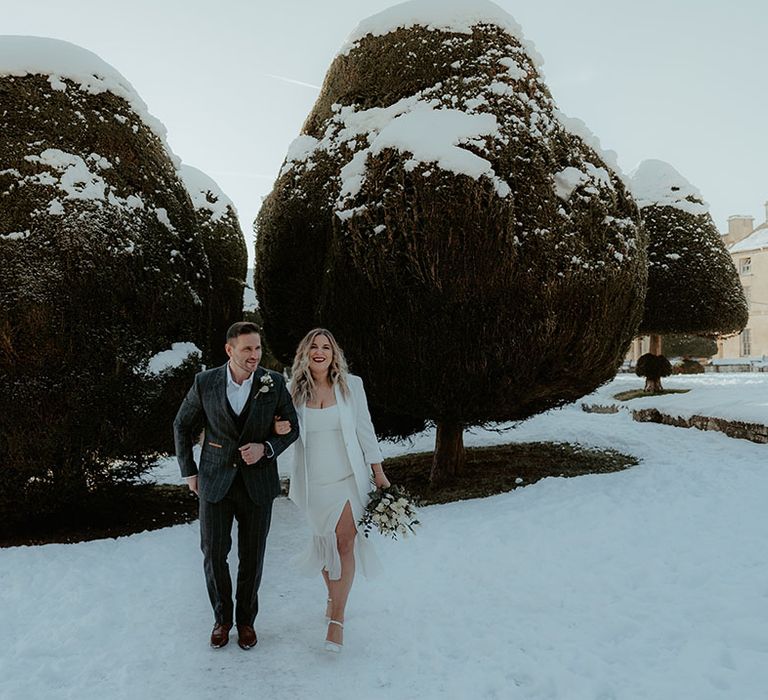 Groom in white shirt with no tie in checkered suit walking through the snow with the bride in all white outfit with blazer 