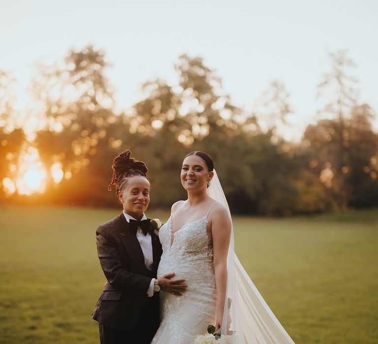 Bride in lace sleeveless v neck Enzoani wedding dress with tulle finish and church length veil posing with bride in black snakeskin print tuxedo with black bowtie and white rose boutonniere at golden hour at Grittleton House lesbian black-tie wedding 