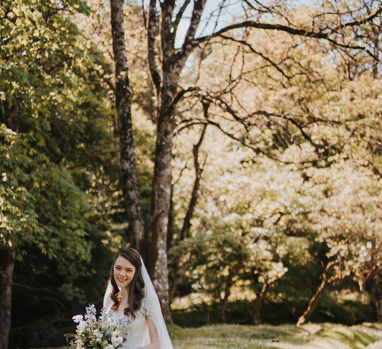 Bride in beaded wedding dress with embroidered cathedral length wedding veil holding white and blue bouquet 