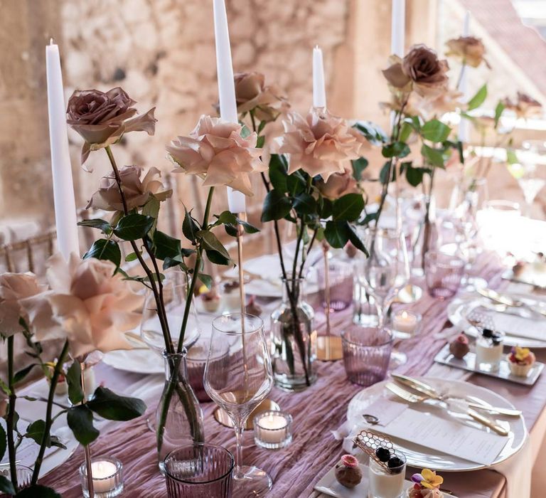 Modern wedding tablescape with lavender tablecloth, lilac tulle table runner, blush garden rose centrepieces in clear vases, tea light candles and a trio of mini desserts 