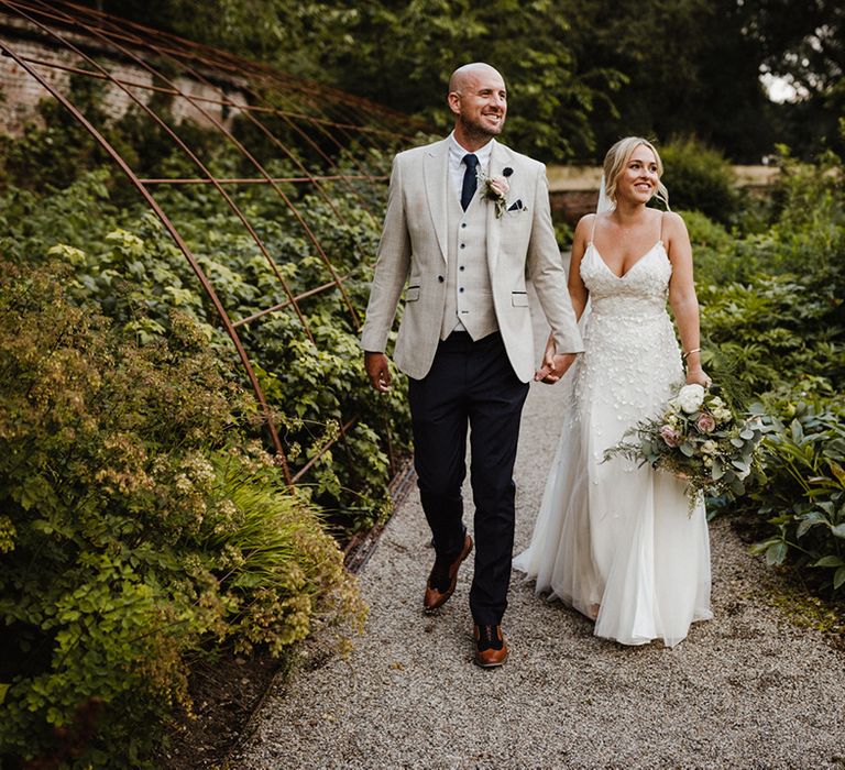 Bride in applique wedding dress by Alexandra Grecco walking along with the groom in a three piece suit with blue tie 