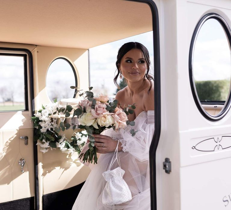 Bride in off the shoulder wedding dress with detachable puff tulle sleeves holding blush garden rose and eucalyptus bridal bouquet sitting in classic vintage wedding car 