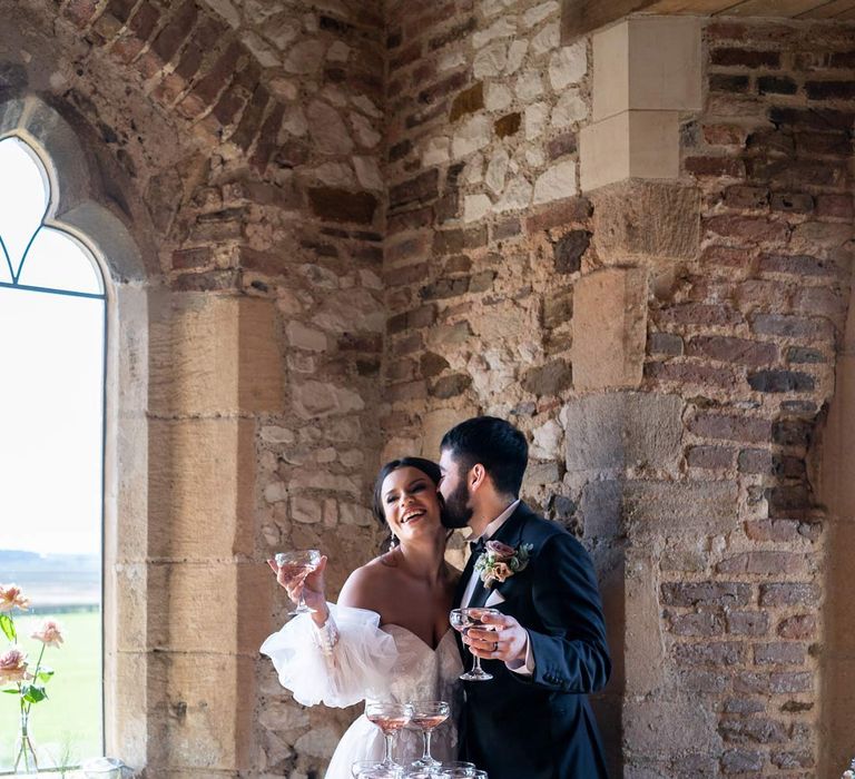 Groom in classic black tuxedo with bowtie and garden rose and eucalyptus boutonniere embracing bride in puddle train ethereal off the shoulder wedding dress with detachable puff tulle sleeves and embroidered rose 3d appliqué details standing by pink champagne tower 