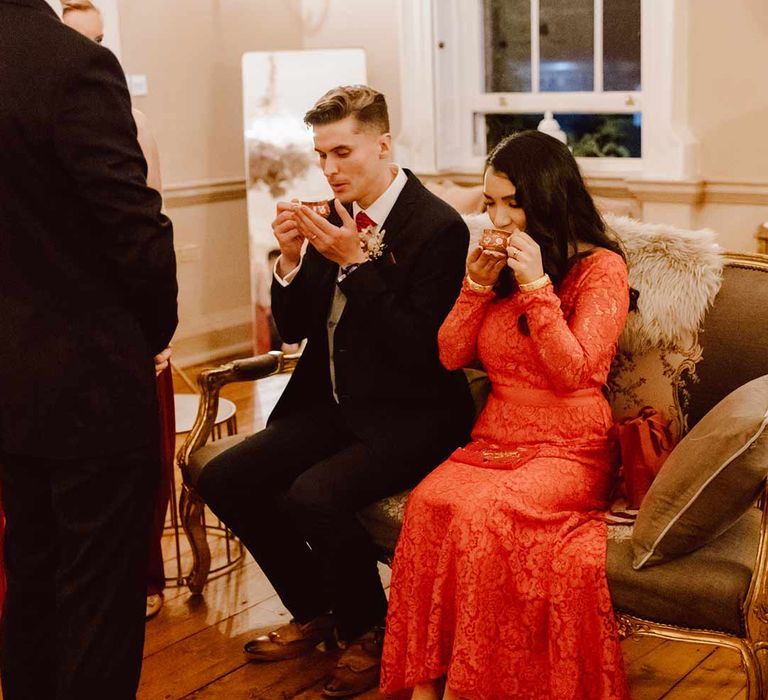 Bride in red lace wedding dress with long sleeves, red and gold hair accessories and golden closed toe wedding heels sipping Chinese tea with groom in deep blue grooms blazer, grey waistcoat, red tie, red pocket square and mixed dried flower boutonniere at Chinese Tea Ceremony