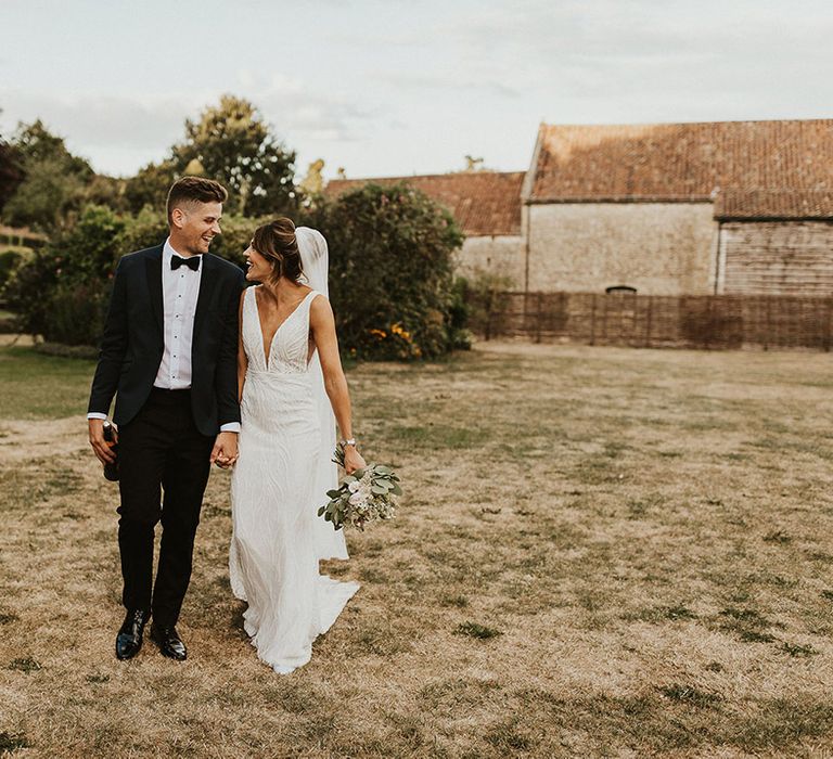 The groom in a navy and black tuxedo walking hand in hand with the bride in a plunging style Made With Love Bridal wedding dress 