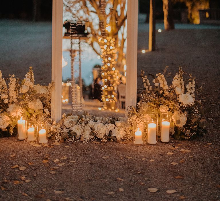 Alternative wedding table plan ideas; large mirror with table plan written on in gold lettering surrounded by foliage, flowers and pillar candles in glass jar candle holders 