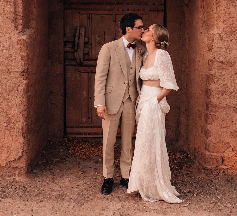 Bride with flowers in her hair wearing a crop top and maxi skirt bridal two piece with the groom leaning for a kiss 