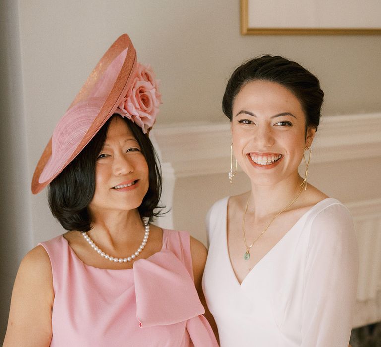 Mother of the bride in pink outfit with fabulous hat smiling with the bride in long sleeve wedding dress