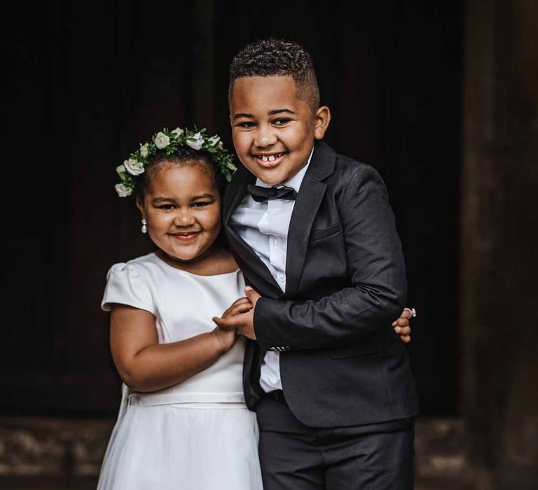 Flower girl wearing short sleeve satin dress and flower crown standing with page boy in black suit and bowtie 